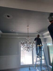 Man on ladder installing chandelier in modern gray room with crown molding.