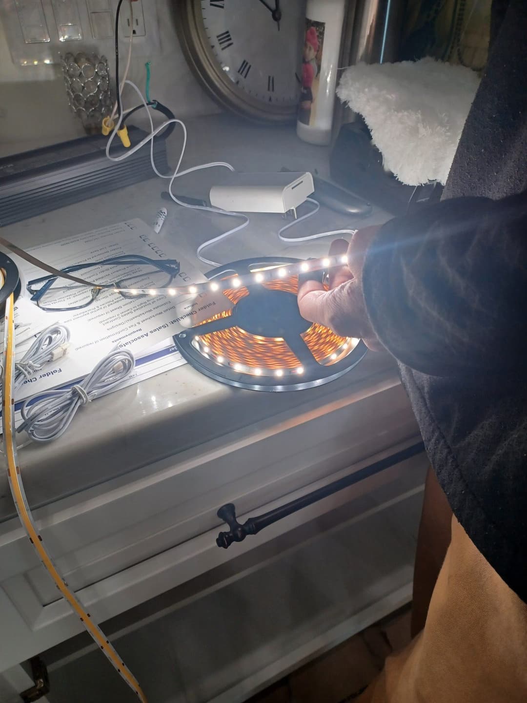 Hand connecting LED strip lights on a countertop with tools and a clock in the background.